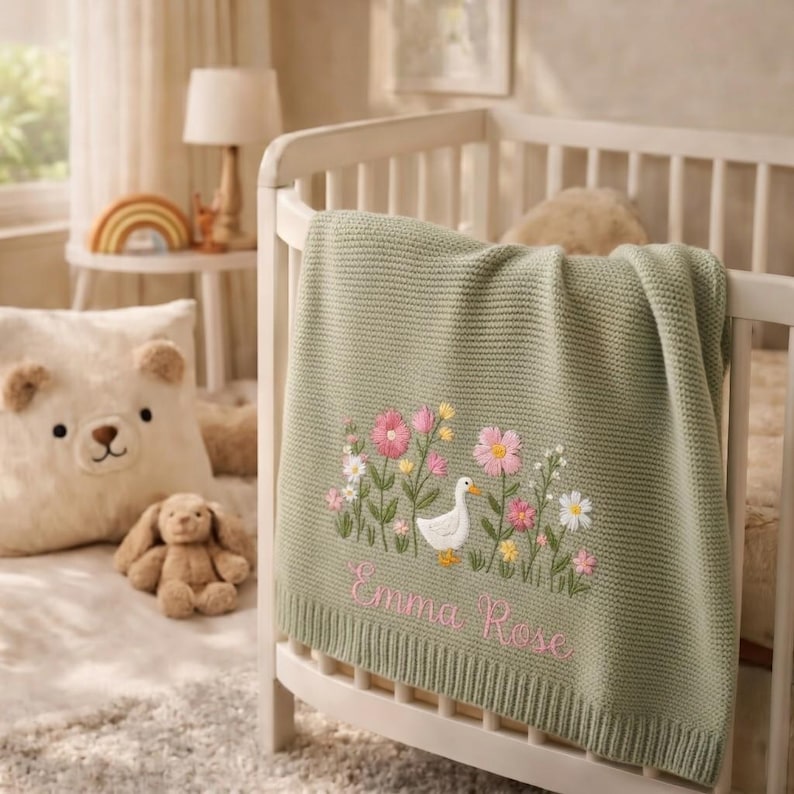 May include: A sage green knitted baby blanket featuring an embroidered scene of a white duck surrounded by pink and yellow wildflowers. The name Emma Rose is embroidered in pink cursive script below the floral design on the textured fabric.