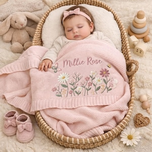 May include: A soft pink knitted baby blanket featuring embroidered wildflowers and a butterfly, with the name Millie Rose stitched in the center. The blanket is draped over a sleeping infant wearing a matching pink headband in a woven basket.