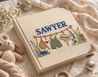 May include: A cream-colored fabric-covered photo album featuring the embroidered name SAWYER in blue. The cover displays an embroidered fishing-themed scene with a net, bobber, overalls, hat, fish, and reeds. It has decorative metal corner accents.