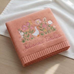 May include: A folded peach-colored knit blanket featuring an embroidered design of a white goose surrounded by various pink, yellow, and white flowers. The name "Julianna Emilia" is embroidered in a cursive script below the floral goose motif.