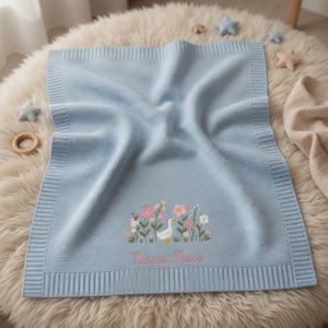 May include: A light blue knitted blanket featuring a ribbed border and a central embroidered design of a white goose surrounded by pink and yellow flowers. The name Emma Rose is embroidered in pink script lettering below the floral illustration.