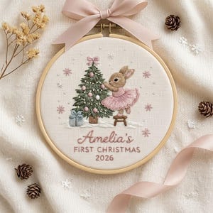 May include: A round wooden embroidery hoop containing fabric with a stitched design of a rabbit in a pink tutu decorating a Christmas tree. The text reads Amelia's FIRST CHRISTMAS 2026. A light pink ribbon is tied to the top of the hoop.