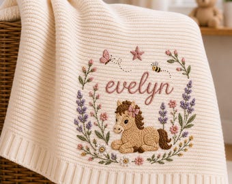 May include: A cream-colored knit blanket featuring an embroidered design of a brown pony, the name evelyn in cursive script, a butterfly, a bee, a star, and a floral wreath with pink, purple, and white flowers and green leaves.