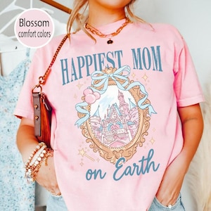 Puede incluir: Camiseta rosa claro con las palabras "Happiest Mom on Earth" en escritura azul. La camiseta presenta una ilustraci&oacute;n enmarcada de un castillo con una cinta azul y una varita m&aacute;gica. Las palabras "Blossom comfort colors" est&aacute;n en un c&iacute;rculo blanco.