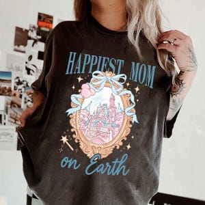 Puede incluir: Camiseta gris oscuro con el texto "Happiest Mom on Earth" en azul claro. La camiseta presenta una ilustraci&oacute;n detallada de un castillo dentro de un marco adornado, con detalles en rosa y azul.
