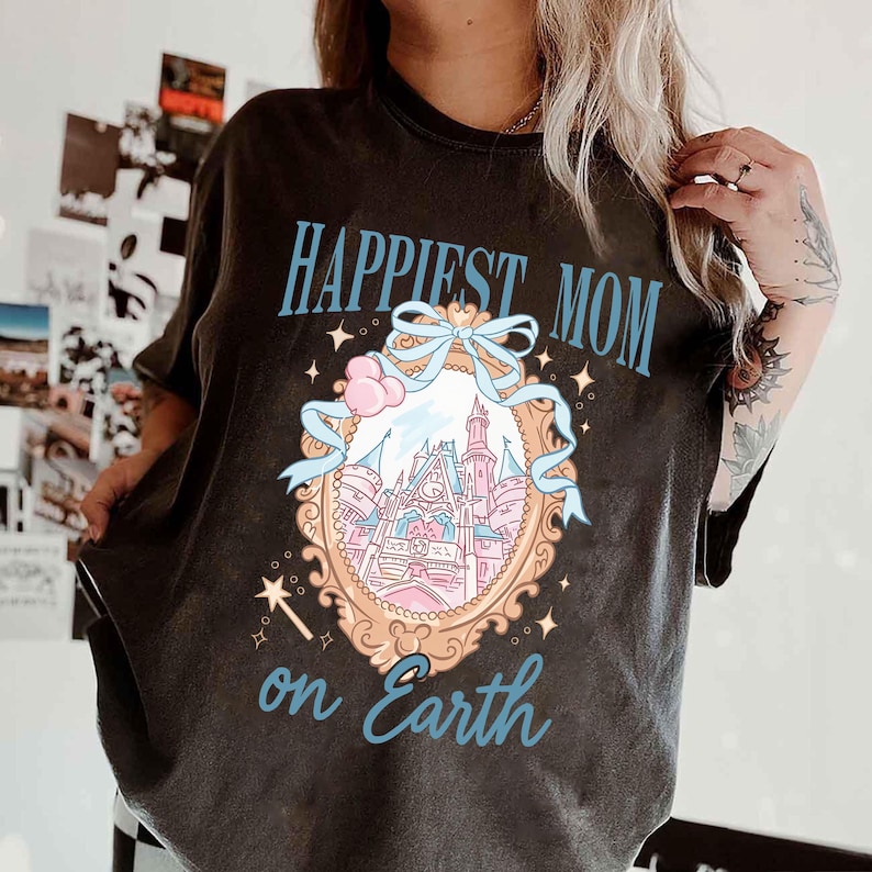 Puede incluir: Camiseta gris oscuro con el texto "Happiest Mom on Earth" en azul claro. La camiseta presenta una ilustraci&oacute;n detallada de un castillo dentro de un marco adornado, con detalles en rosa y azul.