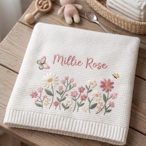 May include: A white waffle-knit baby blanket featuring embroidered floral motifs in shades of pink, white, and green. The name Millie Rose is stitched in pink script above the flowers, accompanied by a small butterfly on the left and right sides.