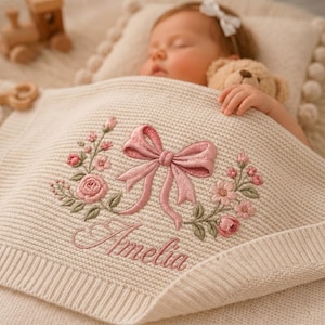 May include: A cream-coloured knit baby blanket featuring an embroidered design of a pink bow centred between two floral sprays. The name Amelia is embroidered in pink cursive script below the bow. The blanket has a ribbed border edge.