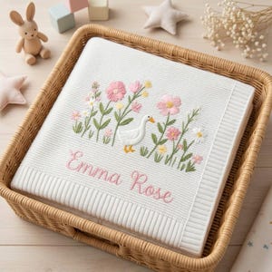 May include: A white knitted baby blanket featuring an embroidered design of a white goose surrounded by pink and yellow flowers with green stems. The name Emma Rose is embroidered in pink cursive script below the floral and goose illustration.