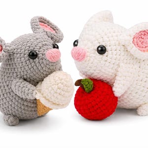 May include: Two crocheted mice, one gray and one white, face each other. The gray mouse holds a crocheted ice cream cone, while the white mouse holds a red crocheted apple. Both have pink ears and noses, and black eyes.