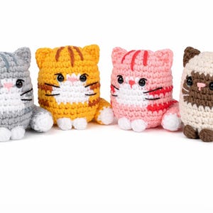 May include: Four handmade crochet cat figurines in various colors: gray, orange, pink, and cream with brown accents. Each cat has a white face and paws, with black eyes and whiskers. The cats are sitting upright.