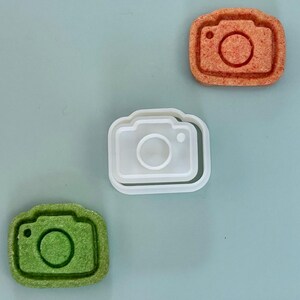 May include: Three items are arranged on a light blue surface: a white plastic camera-shaped cookie cutter, and two cookies. One cookie is orange, the other is green. Both cookies and the cutter have a camera design.