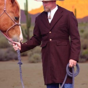 May include: A man in a maroon coat and cowboy hat stands next to a brown horse. The coat has gold buttons and a tailored fit. The man is holding a rope, and the horse is wearing a halter. The background features a desert landscape with cacti and mountains.