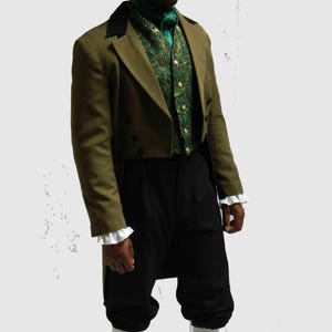 May include: An olive green tailcoat with a black velvet collar, paired with a green paisley vest and black trousers. The outfit includes a green neck scarf and white ruffled cuffs, creating a vintage, formal aesthetic.
