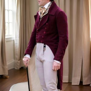 Puede incluir: Un frac burdeos de doble botonadura con botones a juego, combinado con pantalones de color claro y una corbata estampada. El abrigo tiene una cola larga y dividida y se usa con una camisa blanca. Se sostiene un bast&oacute;n negro en la mano.