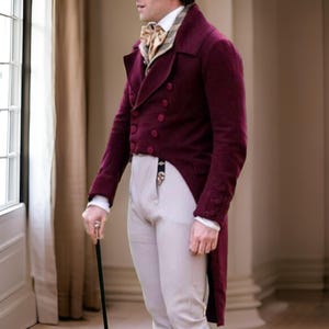 Puede incluir: Un frac burdeos de doble botonadura con botones a juego, combinado con pantalones de color claro y una corbata estampada. El abrigo tiene una cola larga y dividida y se usa con una camisa blanca. Se sostiene un bast&oacute;n negro en la mano.