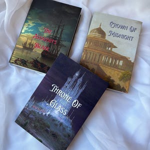 Puede incluir: Tres libros de tapa dura con portadas de temática fantástica están dispuestos sobre una superficie blanca. Los libros se titulan "The Assassin's Blade", "Crown of Midnight" y "Throne of Glass". Las portadas presentan ilustraciones de un barco, un edificio y un castillo.