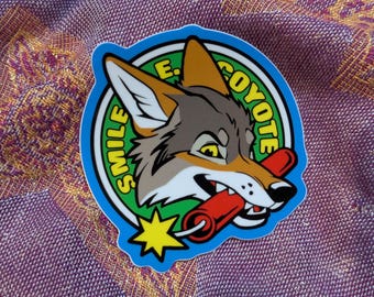 May include: A sticker featuring a cartoon coyote head with the text "SMILE E. COYOTE" in a circular design. The coyote is holding a red firecracker in its mouth, with a yellow starburst graphic.