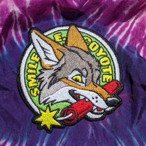Peut inclure: Un patch brodé vert, jaune et blanc avec un coyote de dessin animé tenant un bâton de dynamite rouge. Le patch porte les mots "Smile" et "Coyote" écrits sur le bord.