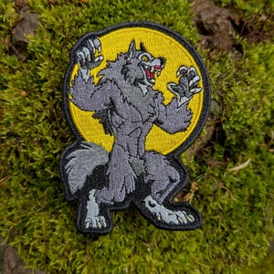 May include: Embroidered patch featuring a werewolf in a menacing pose against a yellow circle. The werewolf is gray with red accents, set against a black border. The patch is placed on a bed of green moss.