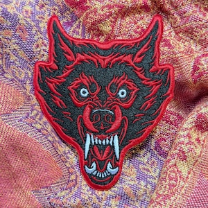 May include: Red and black embroidered wolf head patch with a snarling expression. The wolf's eyes are white and the teeth are white.