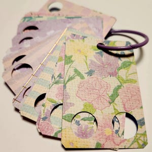 Puede incluir: Un conjunto de etiquetas decorativas unidas a un anillo morado. Las etiquetas presentan varios patrones pastel, incluyendo estampados florales, rayas y diseños a cuadros. Cada etiqueta tiene un recorte circular. La paleta de colores incluye rosa, morado, amarillo y verde.