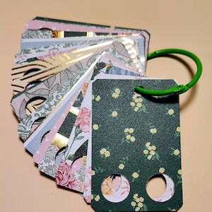 May include: A fan-shaped collection of rectangular paper tags held together by a green metal ring. Each tag features distinct patterns including dark green with yellow flowers, pink roses, gold foil geometric lines, and soft botanical prints.