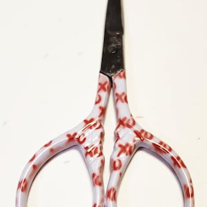 May include: Small scissors with a white handle decorated with a red 'X' pattern. The blades are dark metal. These scissors are likely used for crafts or delicate tasks.