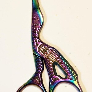 May include: Iridescent embroidery scissors with a crane design. The scissors feature a rainbow finish with a crane-shaped handle and detailed patterns. The blades are sharp and curved, and the finger holes are circular.