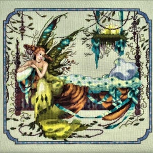 Puede incluir: Diseño de punto de cruz que presenta un hada con alas verdes y marrones, sentada cerca de una cama con una manta con estampado floral. El hada lleva un vestido amarillo y verde. La obra está enmarcada con un borde azul y vides decorativas.