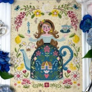 Puede incluir: Obra de arte de punto de cruz que representa a una mujer integrada en un diseño de tetera azul, con motivos florales y de limón. También se incluye una taza de té y una cuchara. El fondo es de color beige claro, enmarcado en blanco.