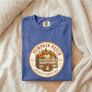 May include: A blue short-sleeve t-shirt featuring a circular graphic with a wooden crate filled with pumpkins. The text reads PUMPKIN PATCH, PROVISIONS CO., EST. 2023, and QUALITY SINCE FALL. The shirt has a Comfort Colors brand label at the neck.