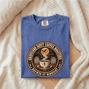 May include: A blue t-shirt featuring a circular graphic with the text "GRAVEYARD SHIFT COFFEE ROASTERS" and "BREWED AT MIDNIGHT". The design depicts a coffee cup with a skull, steam, a full moon, a flying bat, and two tombstones in a dark forest.