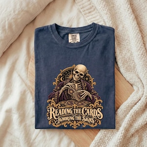 Może przedstawiać: Granatowy t-shirt z grafiką szkieletu w fioletowej szacie trzymającego karty do gry. Wzór zawiera złote ornamenty, elementy niebiańskie oraz napis "READING THE CARDS IGNORING THE SIGNS" wykonany ozdobną czcionką.