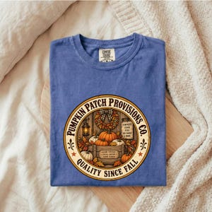 May include: A blue t-shirt featuring a circular graphic with the text Pumpkin Patch Provisions Co. Quality Since Fall. The design includes a wooden crate filled with orange pumpkins, a lantern, a fall wreath, and a sign listing pumpkins, cider, pies, and dry goods.
