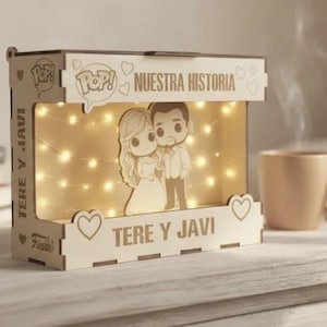Może przedstawiać: Drewniane pudełko świetlne z napisami "Nuestra Historia" i "Tere y Javi" jest podświetlane lampkami choinkowymi. Pudełko przedstawia parę z kreskówek i serca. W pobliżu stoi filiżanka kawy.