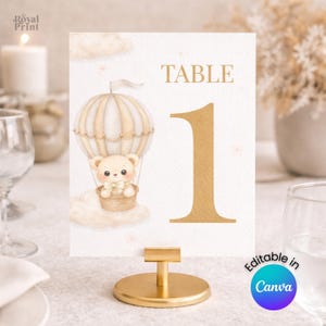 Puede incluir: Una tarjeta con número de mesa con base y soporte dorados. La tarjeta presenta una ilustración de acuarela de un oso de peluche en un globo aerostático, con el número 1 en dorado. El texto "TABLE" está encima del número.