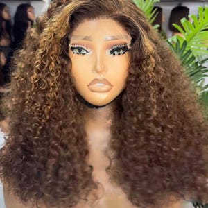 May include: A brown curly wig with highlights is displayed on a mannequin head. The wig features a lace front and a natural-looking hairline. The hair is styled in voluminous, bouncy curls, with lighter brown highlights throughout.