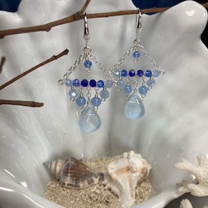 May include: Silver-toned dangle earrings with a geometric design. The earrings feature a chain and are adorned with various shades of blue beads, including teardrop-shaped pendants. The earrings are displayed in a white seashell-shaped bowl.
