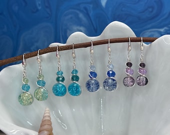 May include: A collection of beaded dangle earrings in various colors, including green, turquoise, and purple. Each earring features multiple round beads wire-wrapped together, suspended from silver-tone ear wires. The earrings are displayed on a branch inside a white seashell.