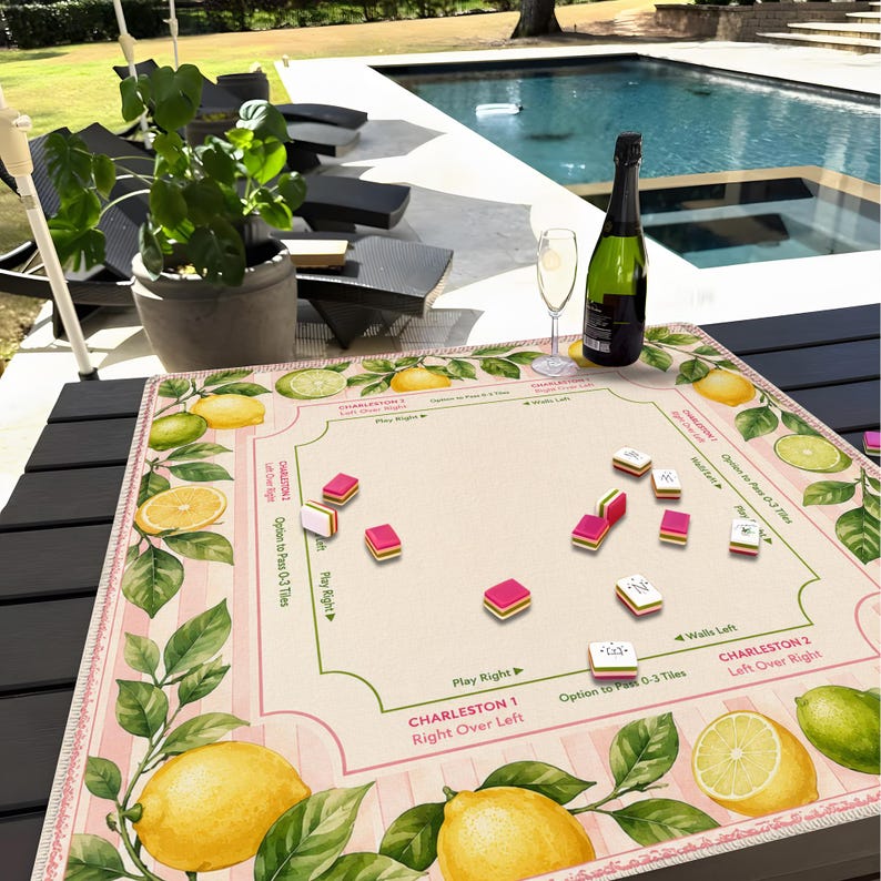 May include: A board game with a pink and cream background, featuring lemons and leaves. The game board has text that reads "Charleston 1" and "Charleston 2". There are game pieces and a bottle of champagne with a glass.