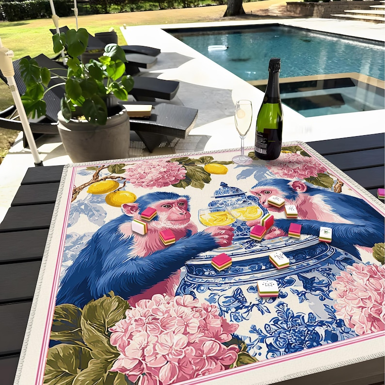 Pu&ograve; includere: Un tappetino da tavolo rettangolare decorativo con due scimmie blu che giocano. Il tappetino presenta un motivo floreale con accenti rosa e blu. Una bottiglia di champagne e un bicchiere sono sul tavolo.