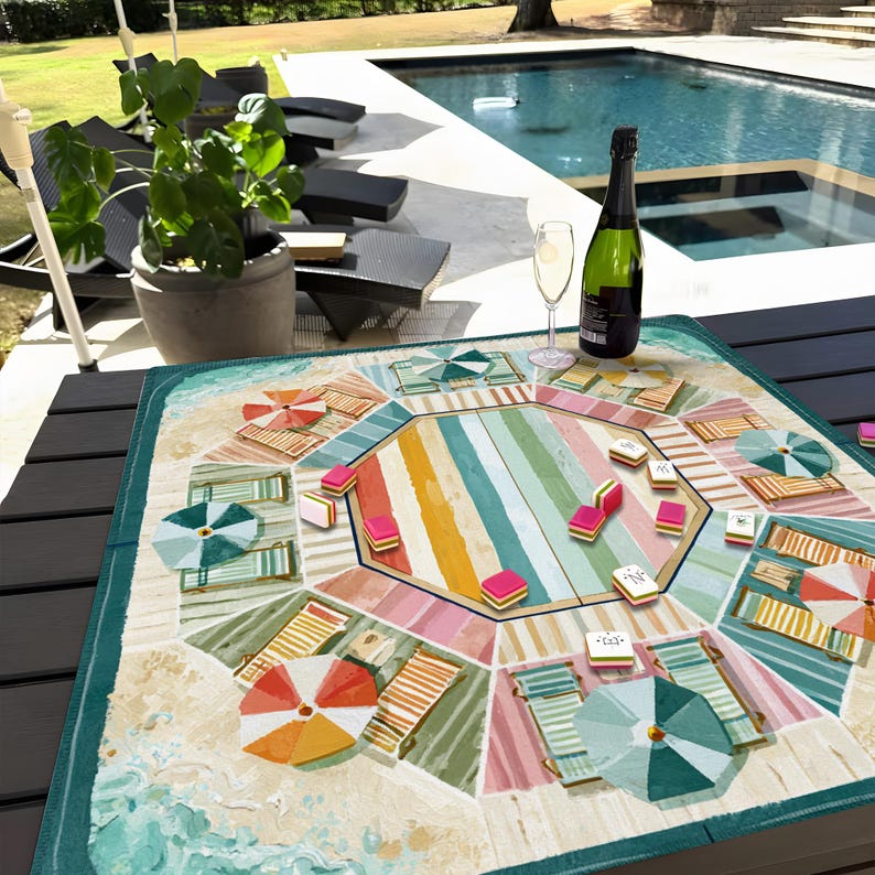 May include: A colorful beach-themed board game with a striped center and illustrations of beach umbrellas, lounge chairs, and sand. A bottle of champagne and a glass of champagne are on the table. The game is set on a dark table near a pool.