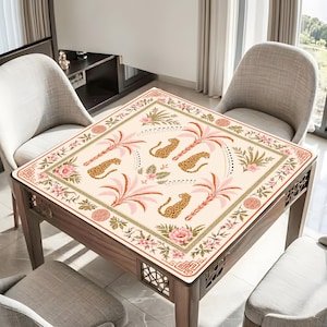 May include: A square table cover featuring a cream-coloured background with a repeating pattern of spotted leopards, pink palm trees, and floral motifs. The border includes intricate geometric designs and circular emblems in shades of pink and green.