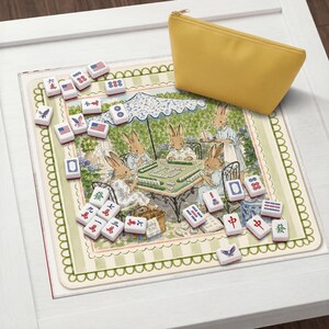 May include: A mahjong set featuring white tiles with red, blue, and black symbols, including American flag designs, arranged on a square mat decorated with an illustration of rabbits playing a game under a parasol, accompanied by a yellow pouch.