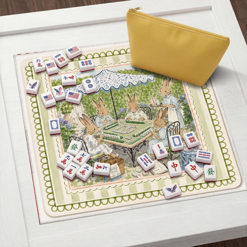 May include: A mahjong set featuring white tiles with red, blue, and black symbols, including American flag designs, arranged on a square mat decorated with an illustration of rabbits playing a game under a parasol, accompanied by a yellow pouch.