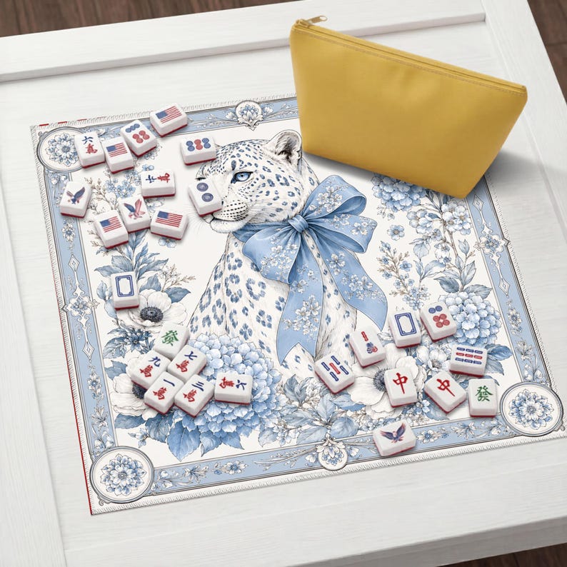 May include: A square mahjong mat featuring a white leopard with a blue floral bow, surrounded by blue and white floral patterns and scattered mahjong tiles with American flag designs and Chinese characters, accompanied by a solid yellow zippered pouch.