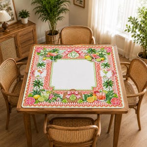 Op de afbeelding: Een vierkante placemat met een tropisch ontwerp met cocktails, fruit en bloemelementen. De mat heeft een wit midden met een decoratieve rand in roze en groen. De tafel is omringd door rotan stoelen.