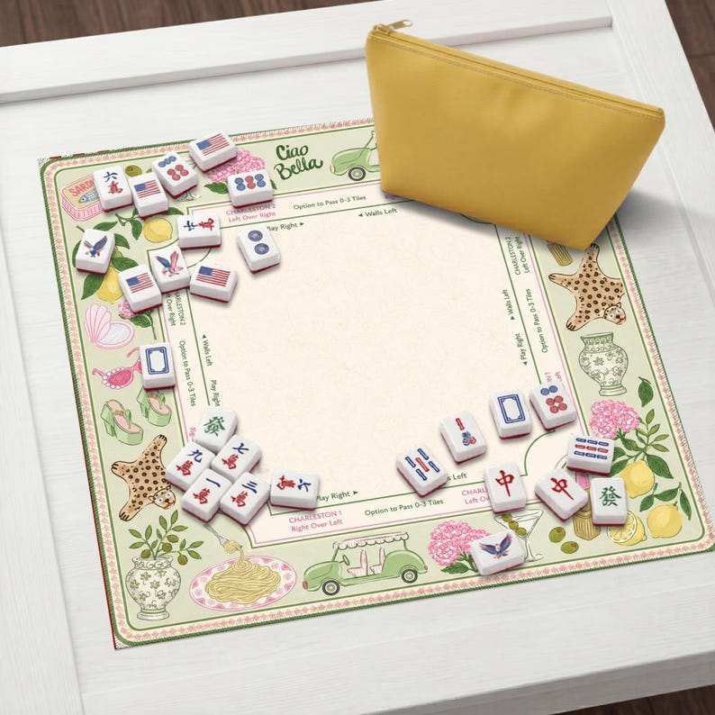 Pu&ograve; includere: Un astuccio giallo con cerniera &egrave; appoggiato su un tappetino decorativo per il gioco del Mahjong. Il tappetino presenta un design floreale e a tema alimentare con le parole "Ciao Bella" e varie tessere del Mahjong. Le tessere del gioco sono bianche con segni rossi e blu.