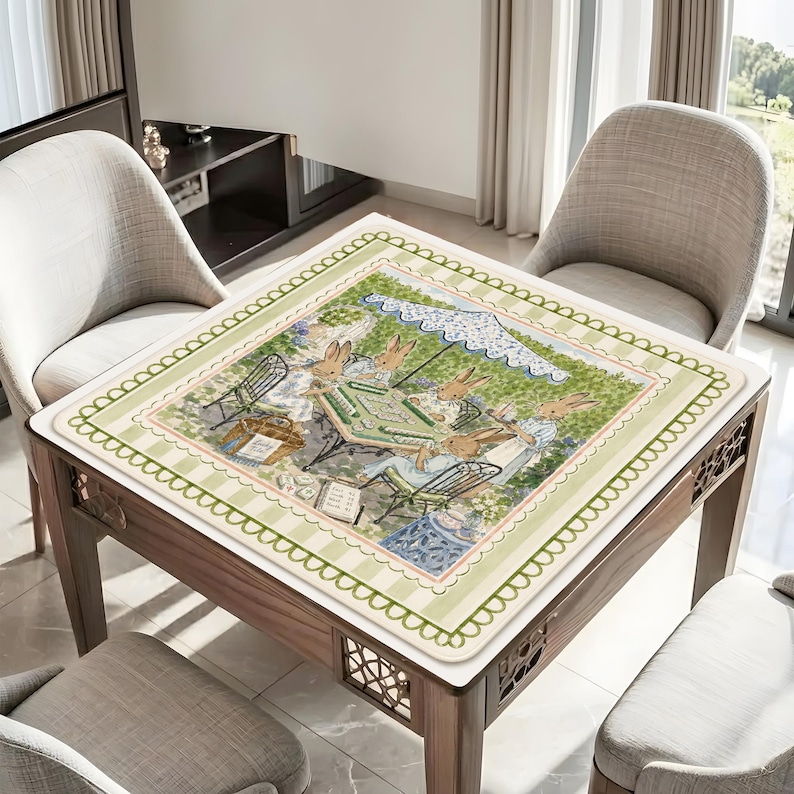 May include: A square table cover featuring a printed illustration of rabbits playing a board game outdoors under a blue and white umbrella. The border has a light green and white striped pattern with a scalloped edge and a decorative green vine motif.
